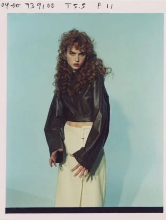 Model with long curly red hair wearing a dark leather top and white skirt against a blue studio backdrop.