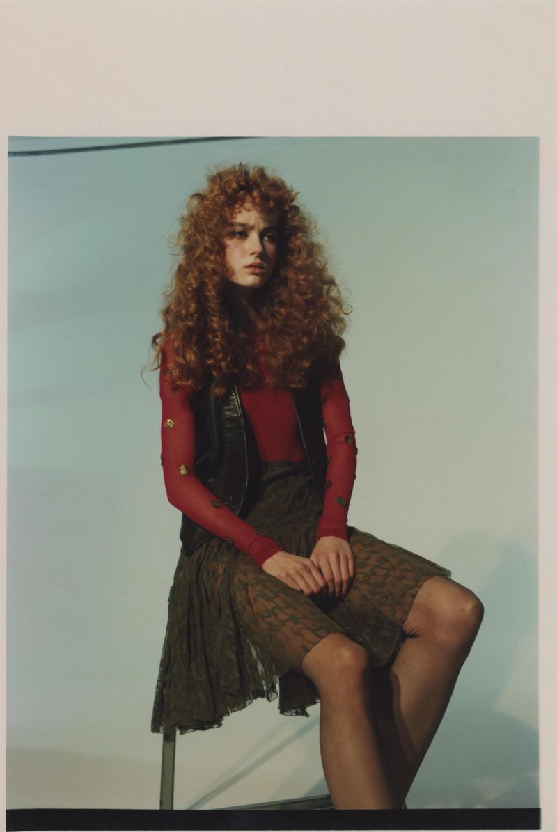 A stylish woman with curly red hair poses on a stool, wearing a red top, vest, and patterned skirt against a light blue background.