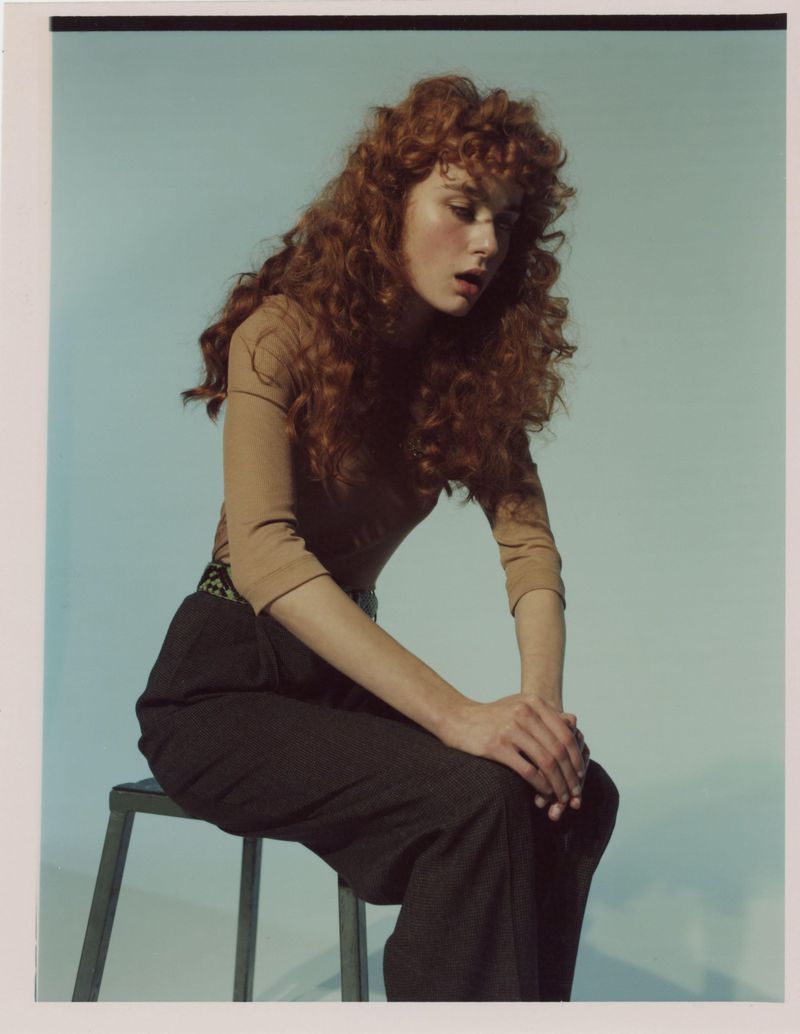 Portrait of a young woman with curly red hair, wearing a tan top and dark pants, sitting on a metal stool against a light blue backdrop.
