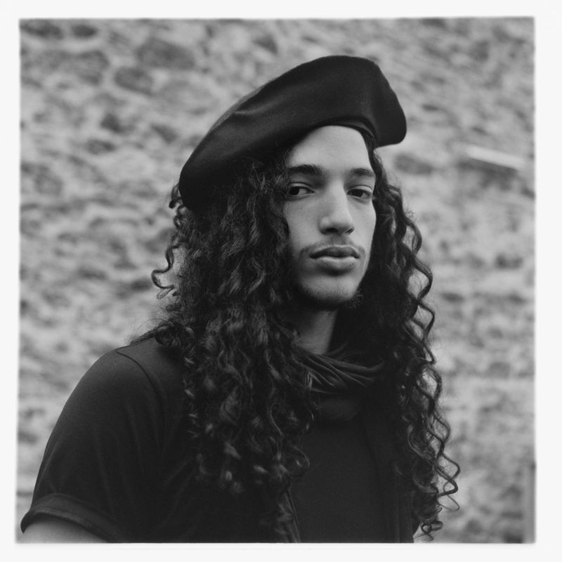Black and white portrait of a young adult with long curly hair, wearing a beret, against a stone wall background.