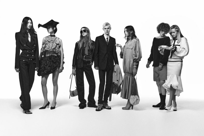 A group of seven diverse models posing together against a stark white studio background, wearing a variety of fashionable designer outfits.