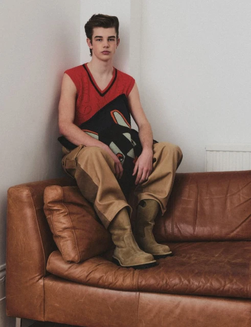 A young adult male is seated on a brown leather couch indoors, wearing a red sweater vest and khaki pants, holding a graphic pillow.