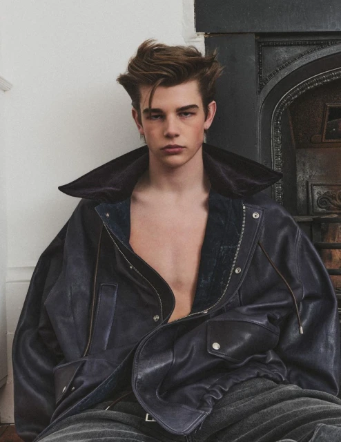 An adult male poses indoors wearing a blue leather jacket. He has brown hair and is seated with an architectural background.