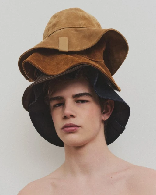 Portrait of a young adult male with fair skin wearing a stack of three bucket hats in brown, tan, and blue against a plain white background.