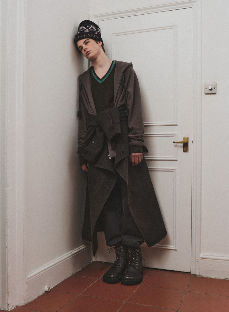 A male model poses indoors, leaning against a white door in a grey-green trench coat, black V-neck sweater, and black patterned beanie.