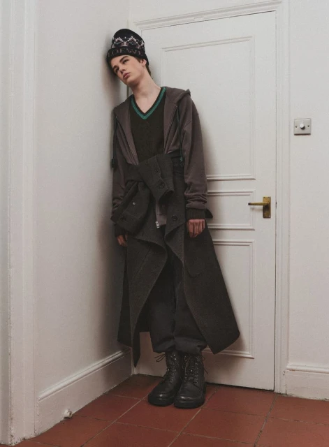 A male model poses indoors, leaning against a white door in a grey-green trench coat, black V-neck sweater, and black patterned beanie.