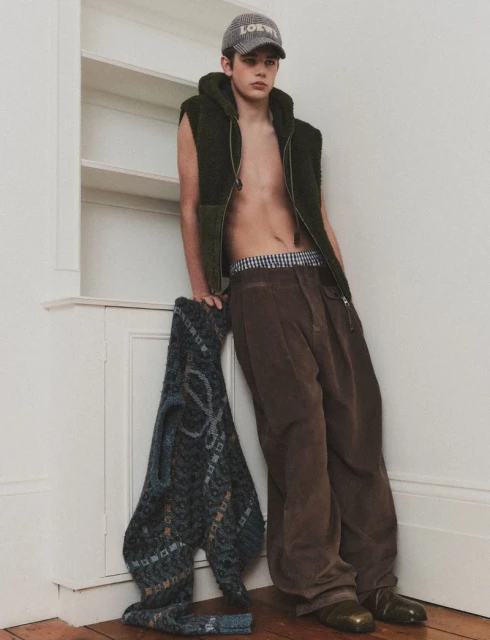 Young man poses indoors wearing a cap, vest, and pants. He leans against a wall with a grey sweater in a casual fashion shot.