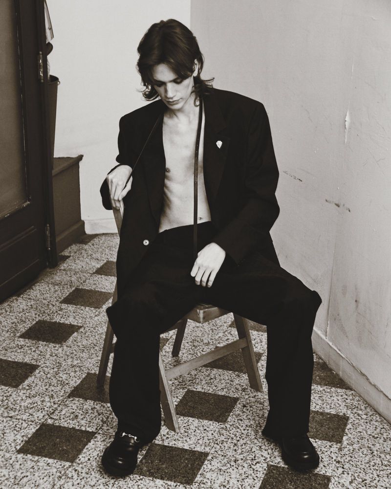 Monochrome portrait of a young male model seated on a wooden chair wearing an oversized black suit jacket and trousers