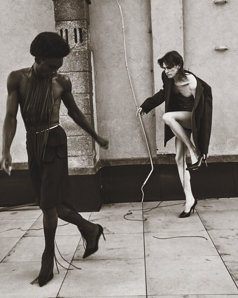 Black and white fashion photograph showcases two slender figures in an outdoor setting, wearing outfits including heels, hose, and a jacket.