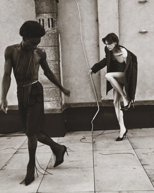 Black and white fashion photograph showcases two slender figures in an outdoor setting, wearing outfits including heels, hose, and a jacket.