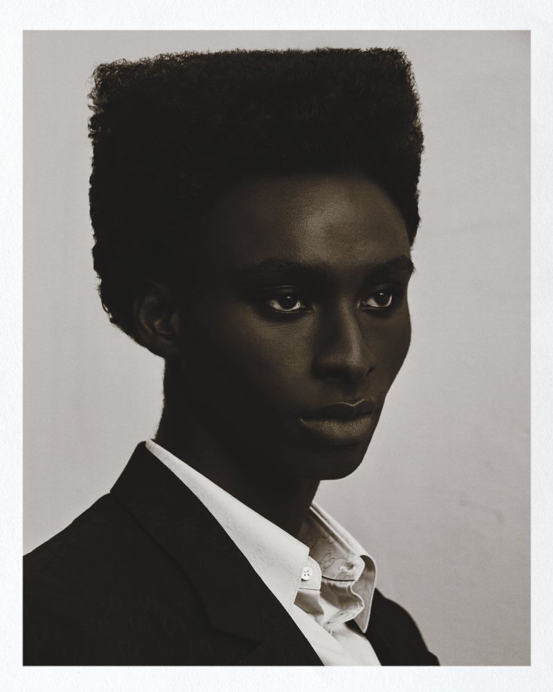 Studio headshot of a Black male model with a high-top fade, wearing a white button-down shirt and black blazer, posing against a neutral backdrop.