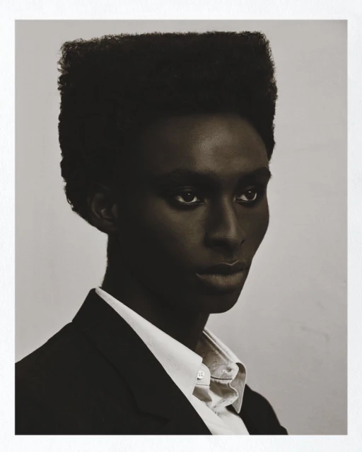 Studio headshot of a Black male model with a high-top fade, wearing a white button-down shirt and black blazer, posing against a neutral backdrop.