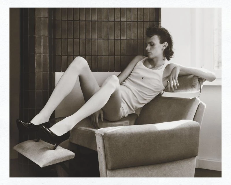 Monochromatic shot of a slender adult in white tank top, shorts, heels, and white tights, sitting on a gray chair with modern background.