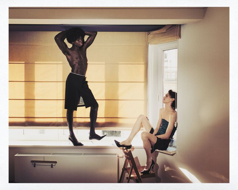 Two people in an indoor setting: a shirtless man in heels stands on a window ledge, while a seated woman looks up at him.