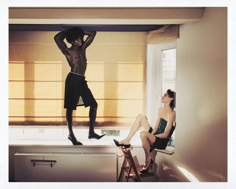 Two people in an indoor setting: a shirtless man in heels stands on a window ledge, while a seated woman looks up at him.