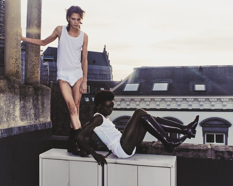 Two fashion models pose on a rooftop wearing white tank tops and briefs with a cityscape in the background during daylight.