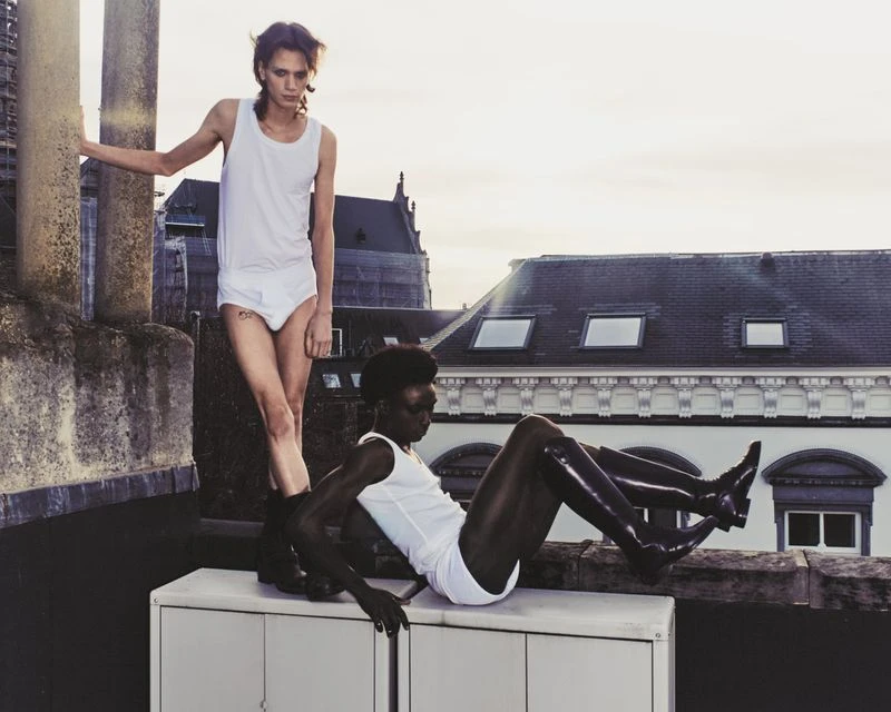 Two fashion models pose on a rooftop wearing white tank tops and briefs with a cityscape in the background during daylight.