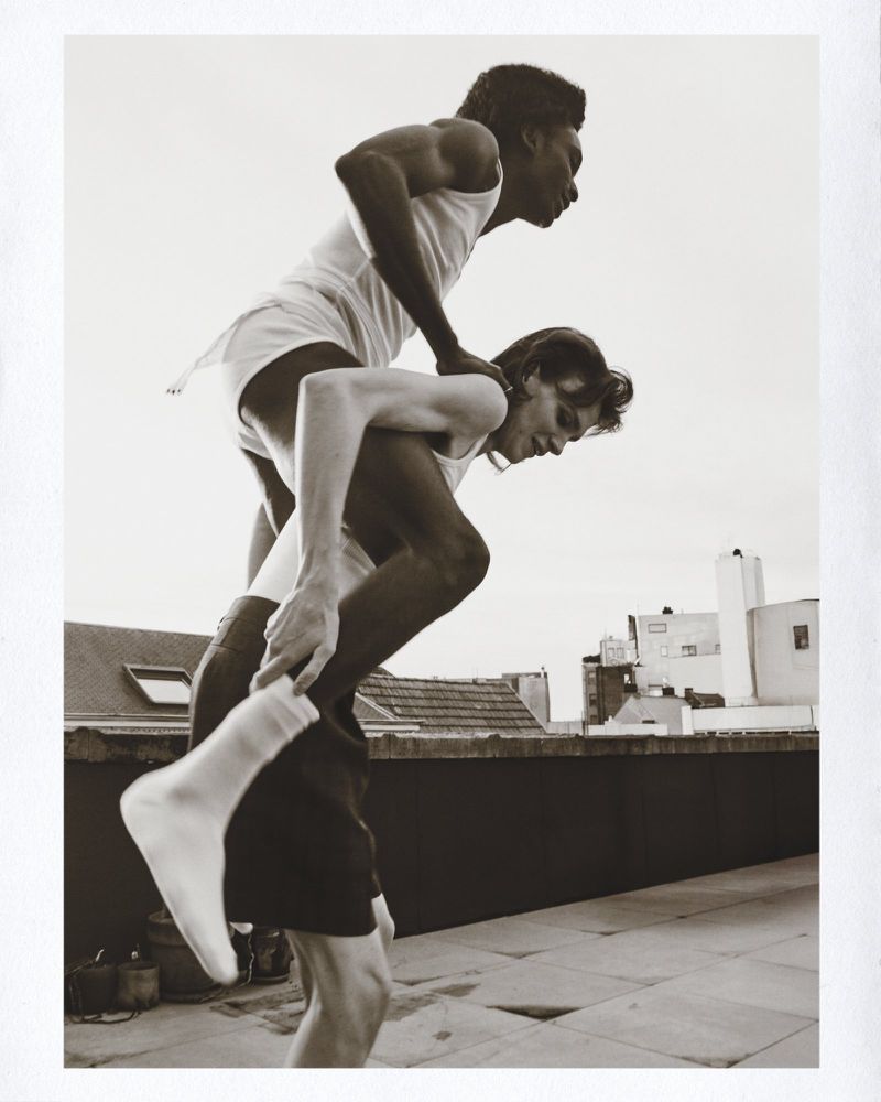 Two men in athletic wear, one giving the other a piggyback ride against an urban skyline in a monochrome photographic style.