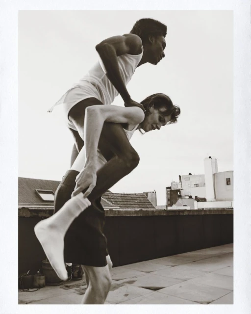 Two men in athletic wear, one giving the other a piggyback ride against an urban skyline in a monochrome photographic style.