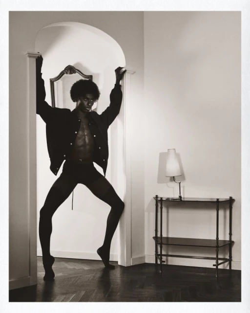 Sepia tone portrait of an adult male with an afro in casual clothes posing in a doorway, in an interior setting with a table.