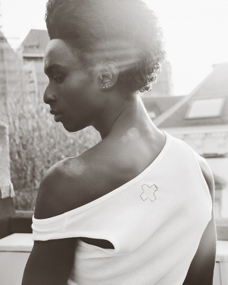 Monochrome portrait of a Black man looking away, wearing a white asymmetrical top with a cross detail, with a building backdrop.