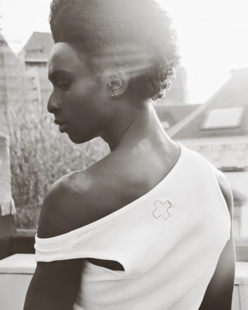 Monochrome portrait of a Black man looking away, wearing a white asymmetrical top with a cross detail, with a building backdrop.