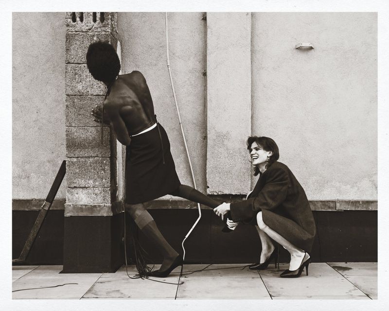 Black and white photo of two women in a playful pose; one woman is leaning on a wall while the other is pulling her leg.
