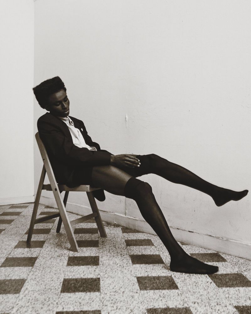 Elegant black model poses on a chair in hosiery, with a slight head tilt, against a minimalist interior backdrop and tiled flooring.