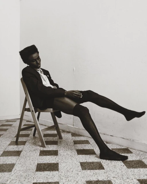 Elegant black model poses on a chair in hosiery, with a slight head tilt, against a minimalist interior backdrop and tiled flooring.