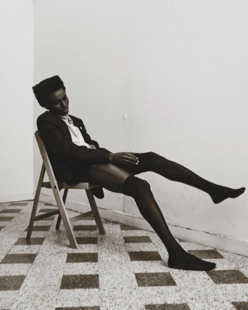 Elegant black model poses on a chair in hosiery, with a slight head tilt, against a minimalist interior backdrop and tiled flooring.