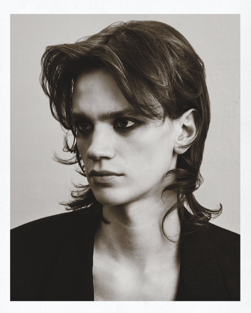 Monochrome portrait of a young adult with medium-length brown hair and dark eye makeup, wearing a black jacket against a neutral background.