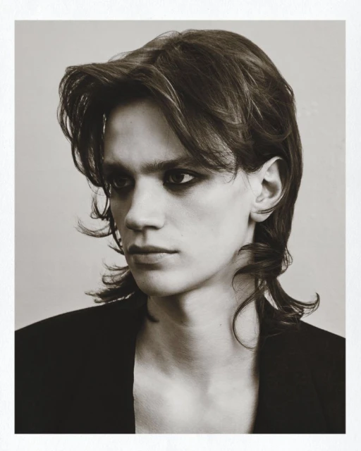 Monochrome portrait of a young adult with medium-length brown hair and dark eye makeup, wearing a black jacket against a neutral background.
