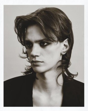 Monochrome portrait of a young adult with medium-length brown hair and dark eye makeup, wearing a black jacket against a neutral background.