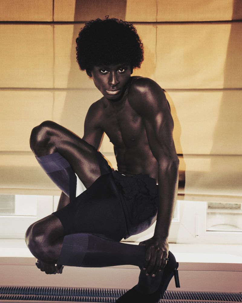Athletic adult male model in squat pose wearing shorts and high heels in an indoor setting with natural light.