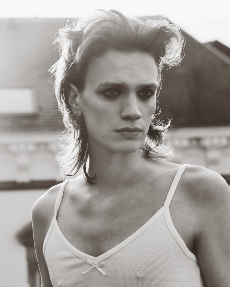A close-up portrait of a person with styled hair and dark eye makeup wearing a white tank top against an outdoor background.