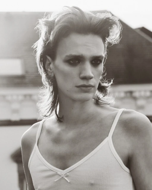 A close-up portrait of a person with styled hair and dark eye makeup wearing a white tank top against an outdoor background.
