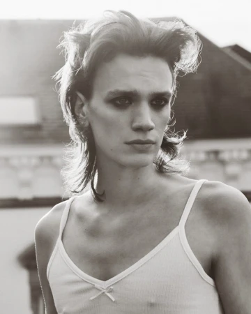 A close-up portrait of a person with styled hair and dark eye makeup wearing a white tank top against an outdoor background.