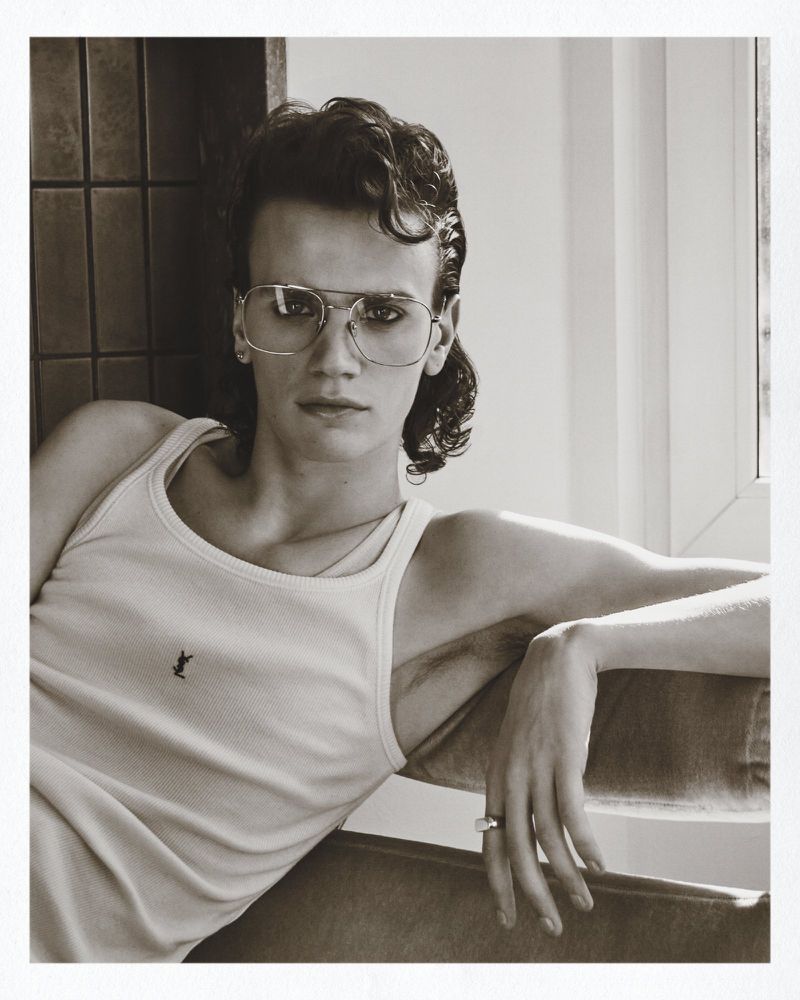 Monochrome portrait of a young man wearing a ribbed tank top and wire-rimmed glasses seated indoors.