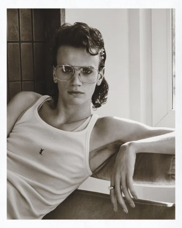 Monochrome portrait of a young man wearing a ribbed tank top and wire-rimmed glasses seated indoors.