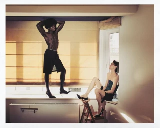 Two male models posing indoors near a window, one standing and one seated on a window sill wearing high heels.