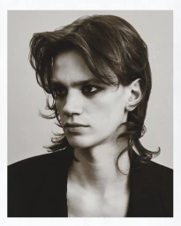 Portrait of a young male model with dark eye makeup and layered dark hair wearing a black jacket against a light neutral background.