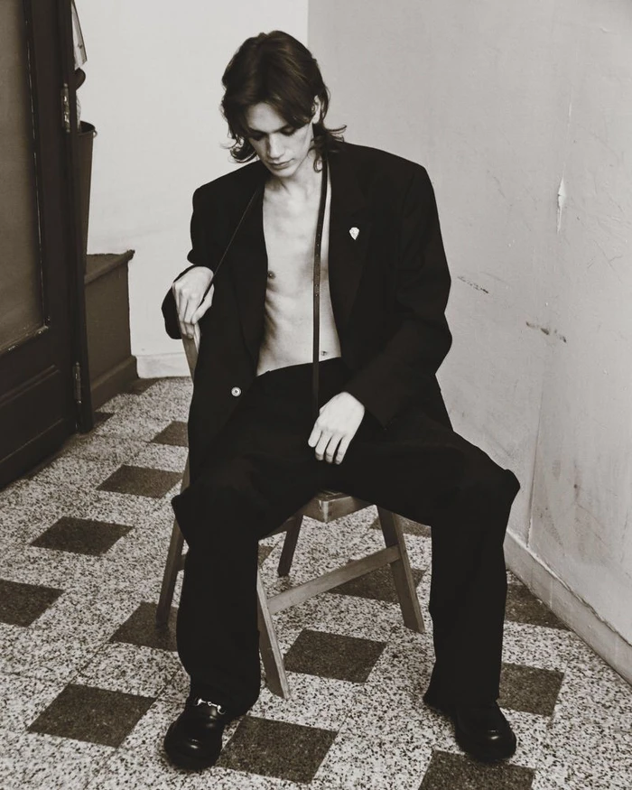 Monochrome portrait of a young male model seated on a wooden chair wearing an oversized black suit jacket and trousers