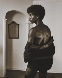 Black man with afro wearing a dark jacket slipping off his shoulders, standing in a room with an arched doorway and a framed mirror.