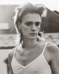 Portrait of a young male model with dark eye makeup and layered hair wearing a white ribbed tank top outdoors.