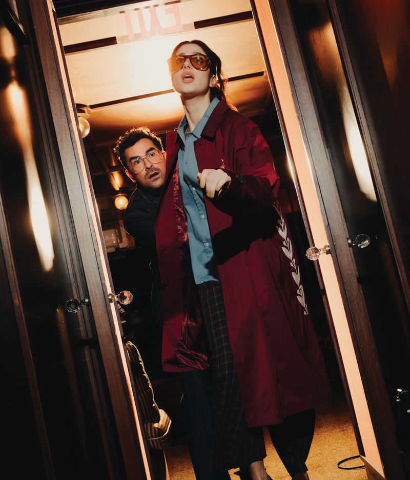 Two people in a doorway with warm lighting: a young adult in a red coat and plaid pants stands next to an adult male peeking out.