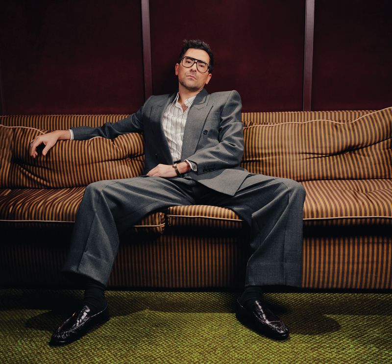 A man in a grey suit sits on a striped couch in a studio, looking at the camera with glasses and a confident expression, indoor setting.