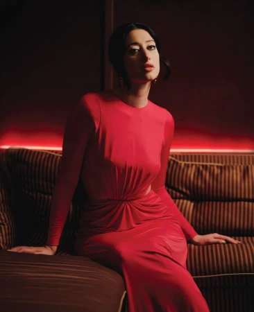 Elegant adult woman with dark hair poses in a long-sleeve red dress while sitting on a striped sofa in a dimly lit indoor setting.