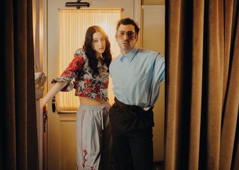 A fashionable couple poses indoors in front of a doorway with curtains, featuring a woman in a floral crop top and a man in sunglasses.