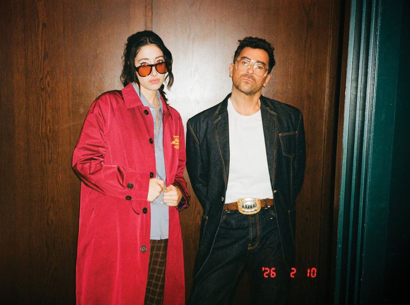 Two people pose indoors against a wooden wall; a woman in a red coat and the man in a denim jacket and white tee.