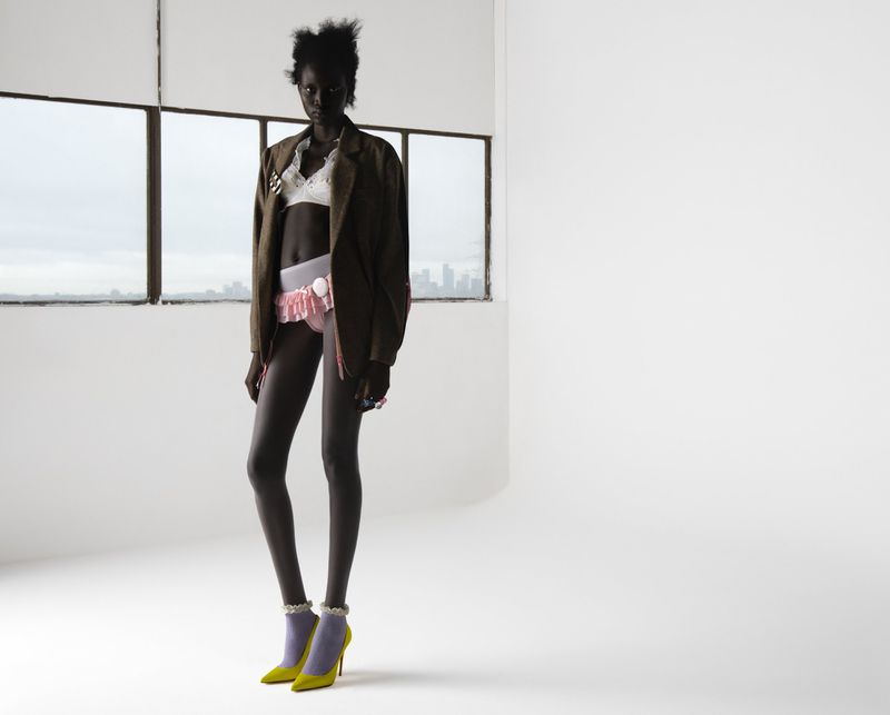 Model Aluel Keror in oversized brown blazer, white bralette, and ruffled shorts standing against a white backdrop.